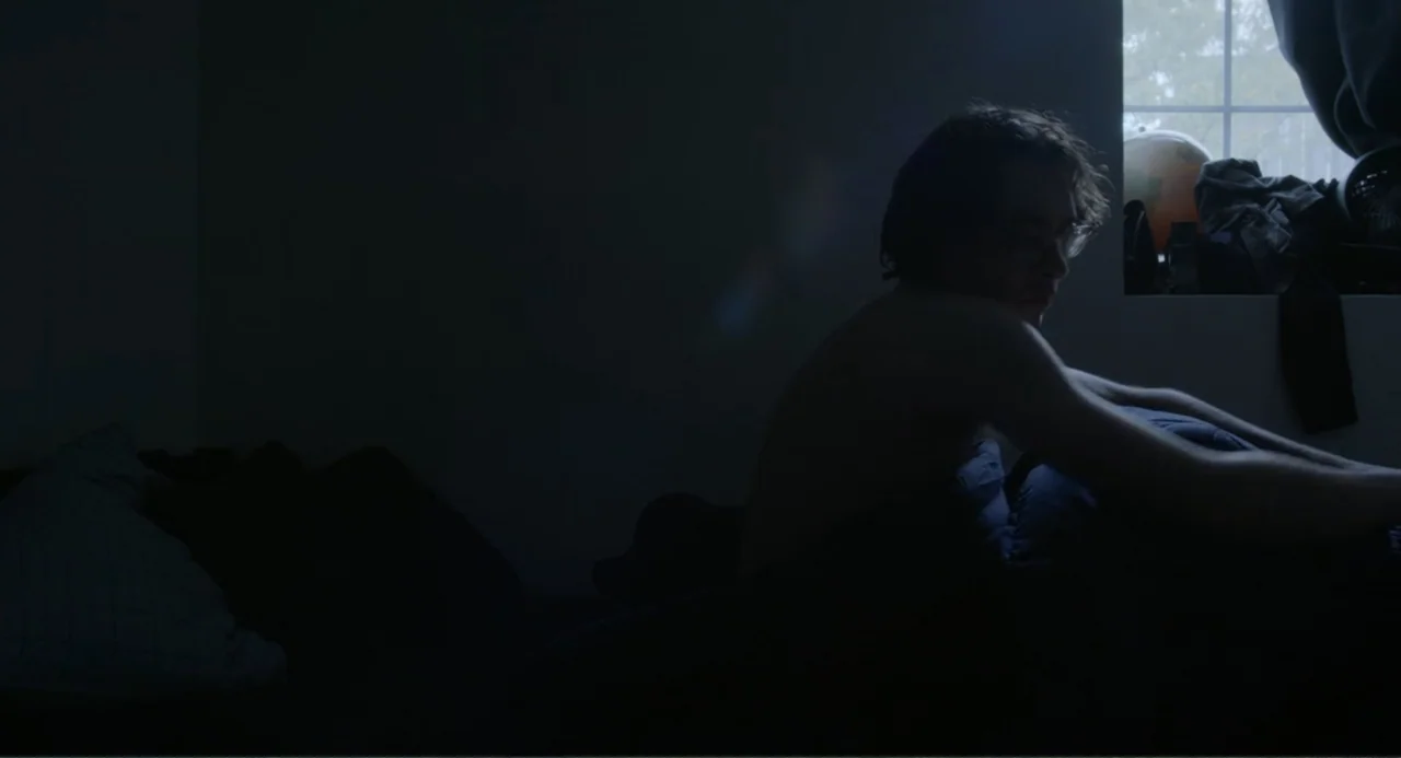 This Closeness (2024) movie still: medium — A dark bedroom interior features a person sitting on a bed near a window. The subject faces toward the…