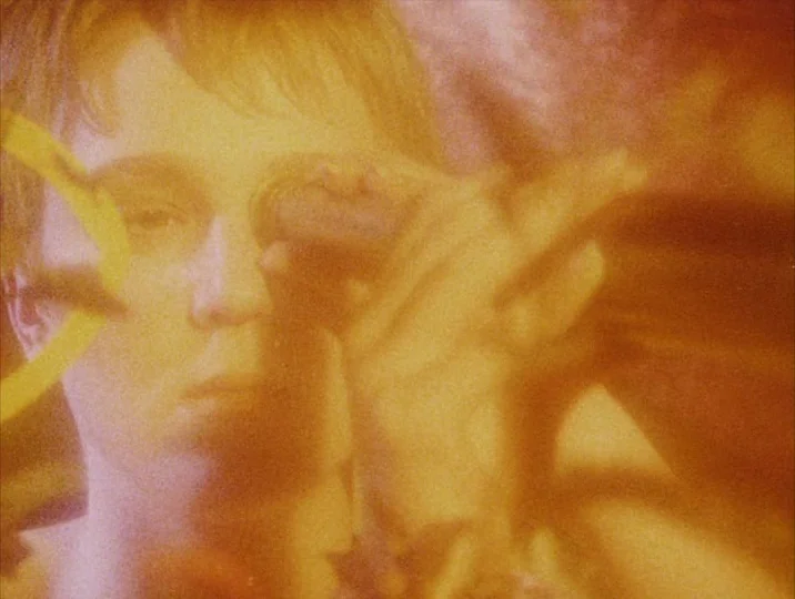 Careful (1992) movie still: closeup — A close-up shot shows a person partially obscured by thick orange lens flares. The figure looks directly into…