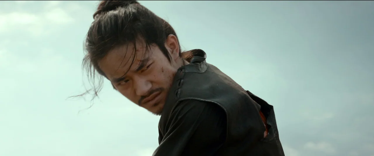 Full River Red (2023) movie still: medium — An open field under a hazy overcast sky during the day. A young man with hair in a topknot looks…