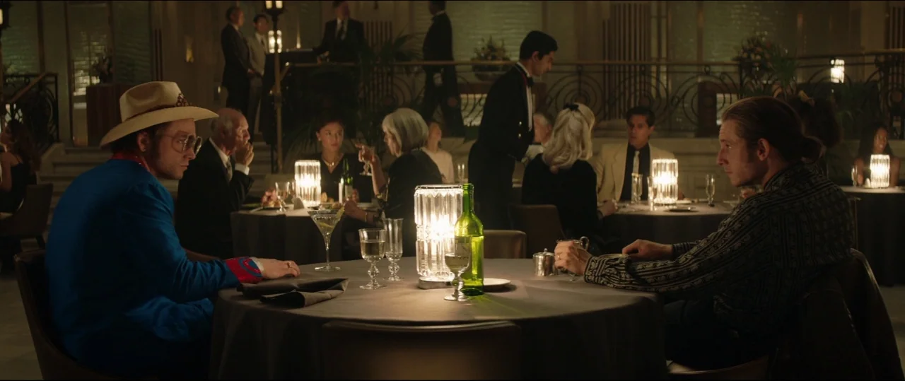 Rocketman (2019) movie still: medium — A high-end restaurant with circular tables and dim lighting in a large room. Two men sit at the foreground…