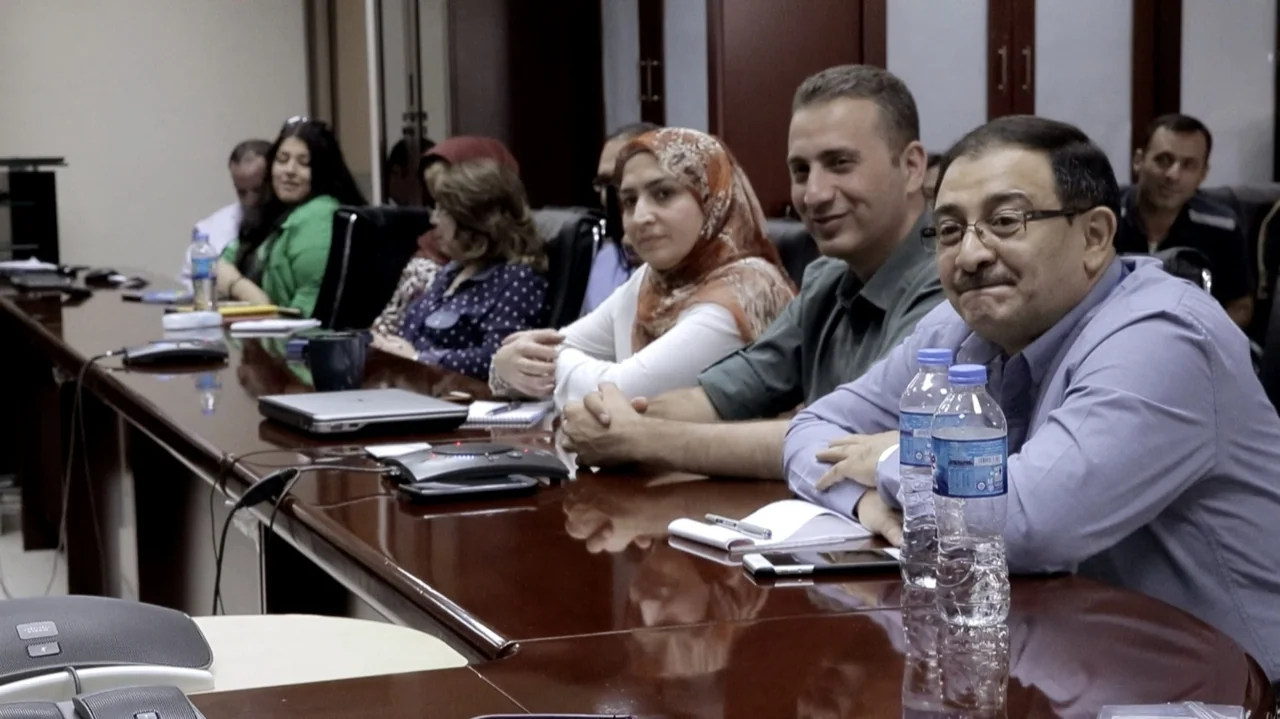 City of Two Springs (2019) movie still: medium — A modern boardroom features a large polished wooden table with assorted attendees present. Several…