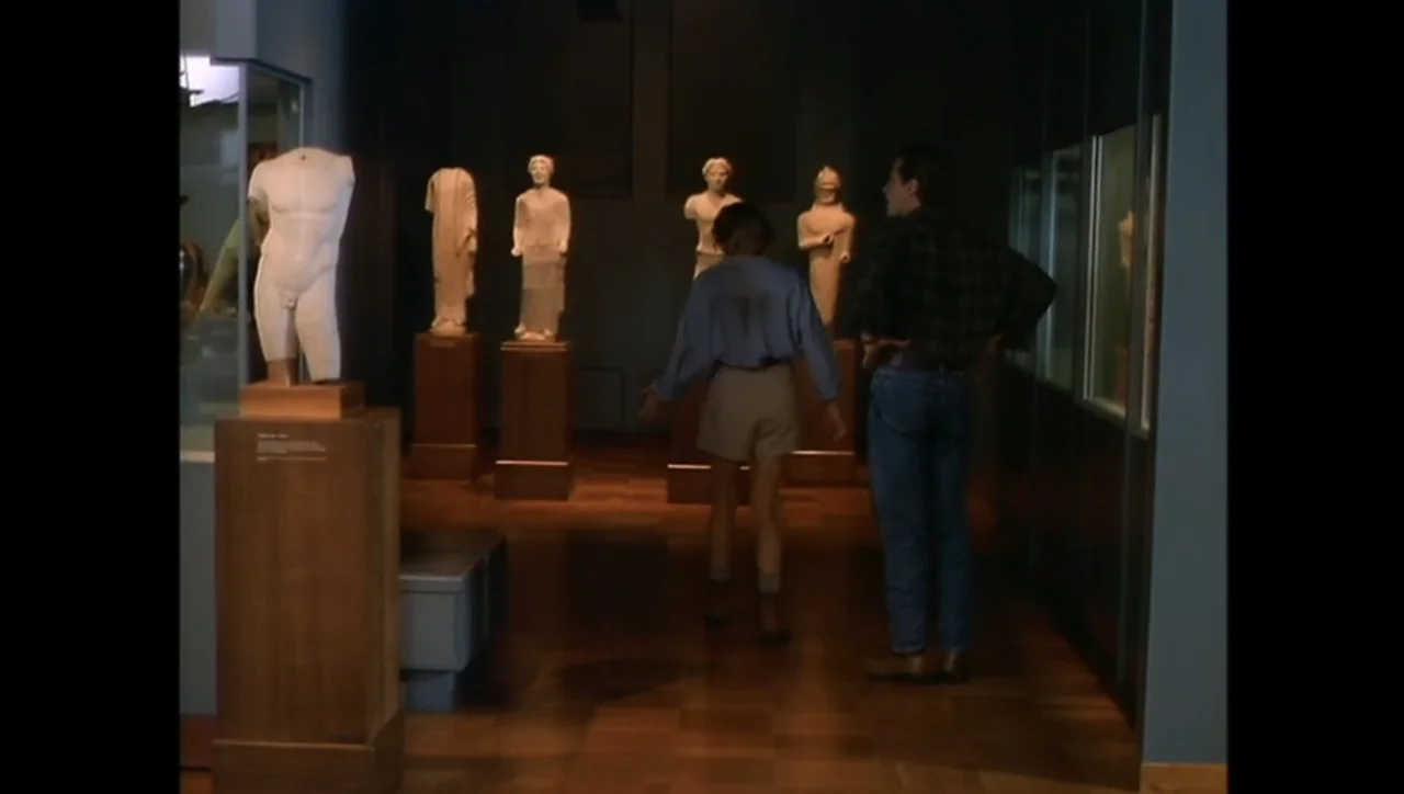 The Awakening (1995) movie still: medium — A dark museum gallery displays several white stone statues on pedestals along a hallway. Two people stand…