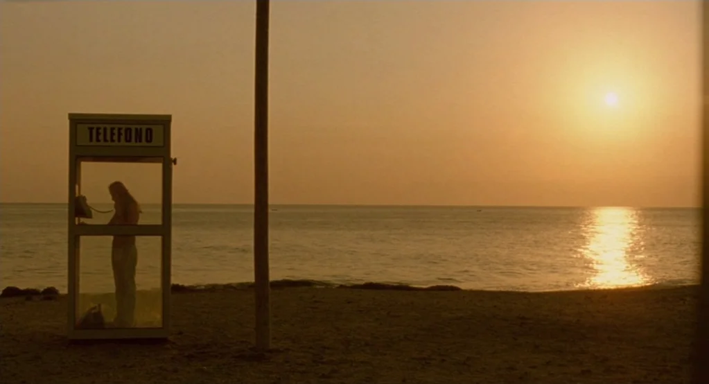 Morvern Callar (2002) movie still: wide — A desolate sandy beach at golden hour features a single glass-enclosed telephone booth. A person stands…