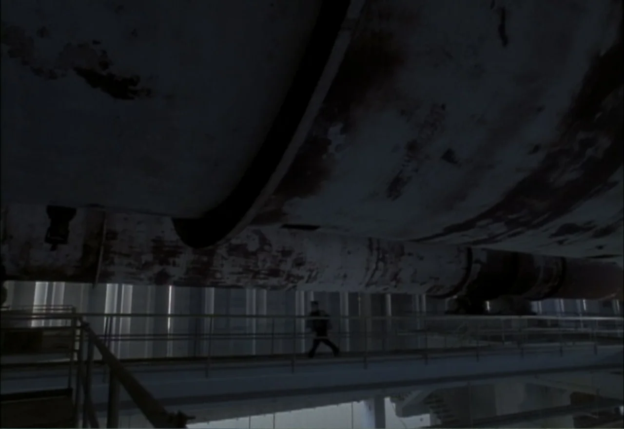 Ambushed (1998) movie still: wide — A vast industrial interior features a massive rusted pipe spanning the upper frame. A single person walks along…