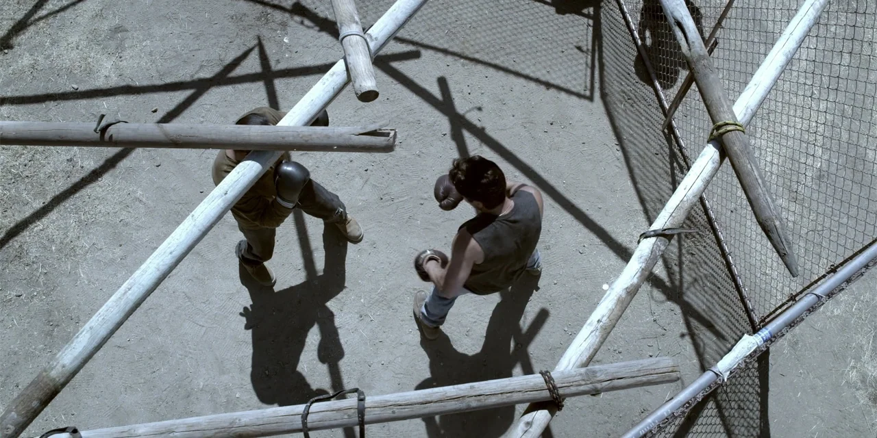 7th & Union (2021) movie still: wide — A makeshift training enclosure constructed from wooden poles and wire fencing on dirt ground. Two men engage…