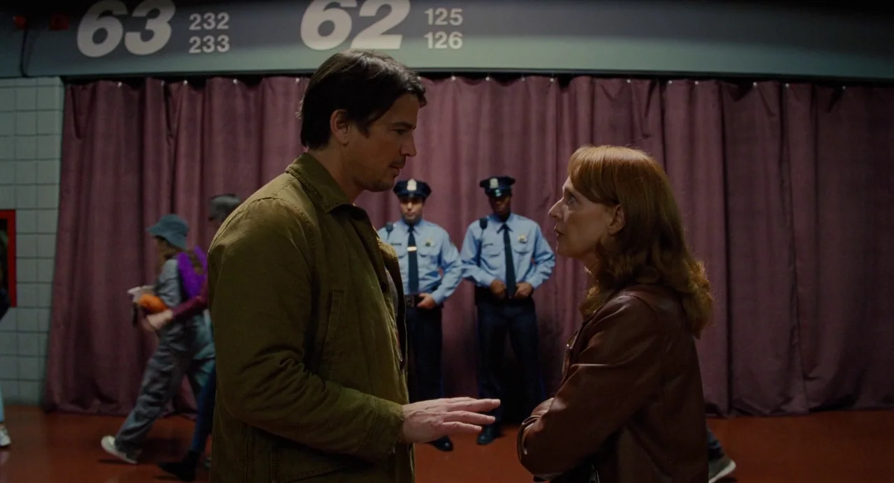 Trap (2024) movie still: medium — A corridor features purple curtains lining the walls with signage indicating numbering sequences above. A man and…