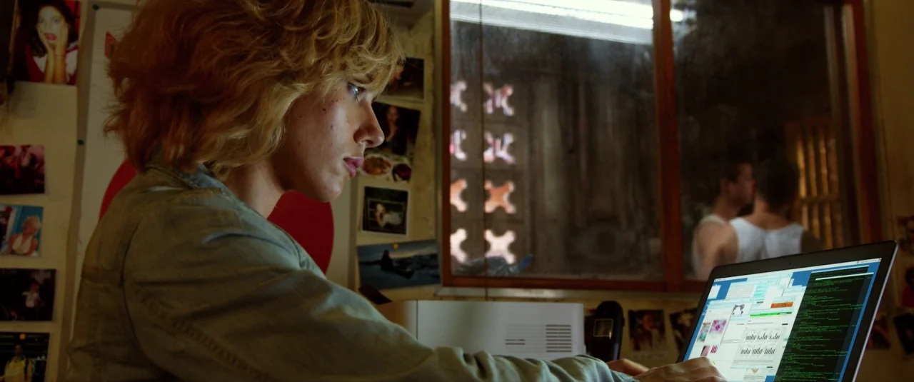 Lucy (2014) movie still: medium — A cluttered room features numerous photos taped to the wall near a window. A blonde woman sits at a desk while…