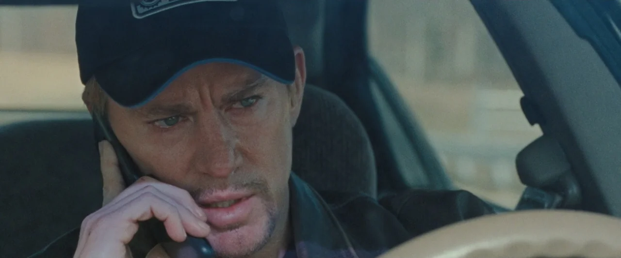 Roofman (2025) movie still: closeup — The interior of a vehicle during daylight hours provides the setting for the shot. A man wearing a baseball…