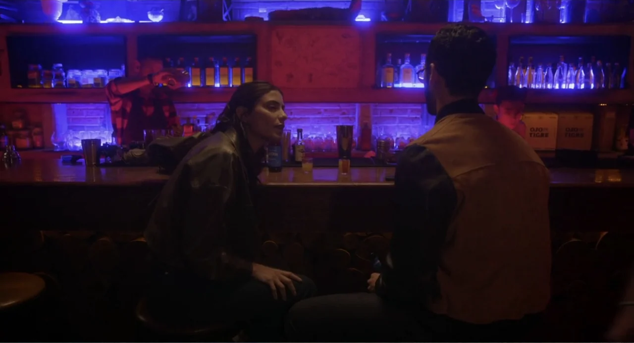 Straight (2024) movie still: medium — A dimly lit bar with neon-illuminated liquor bottles on shelves and wooden decor. A woman and a man sit at the…