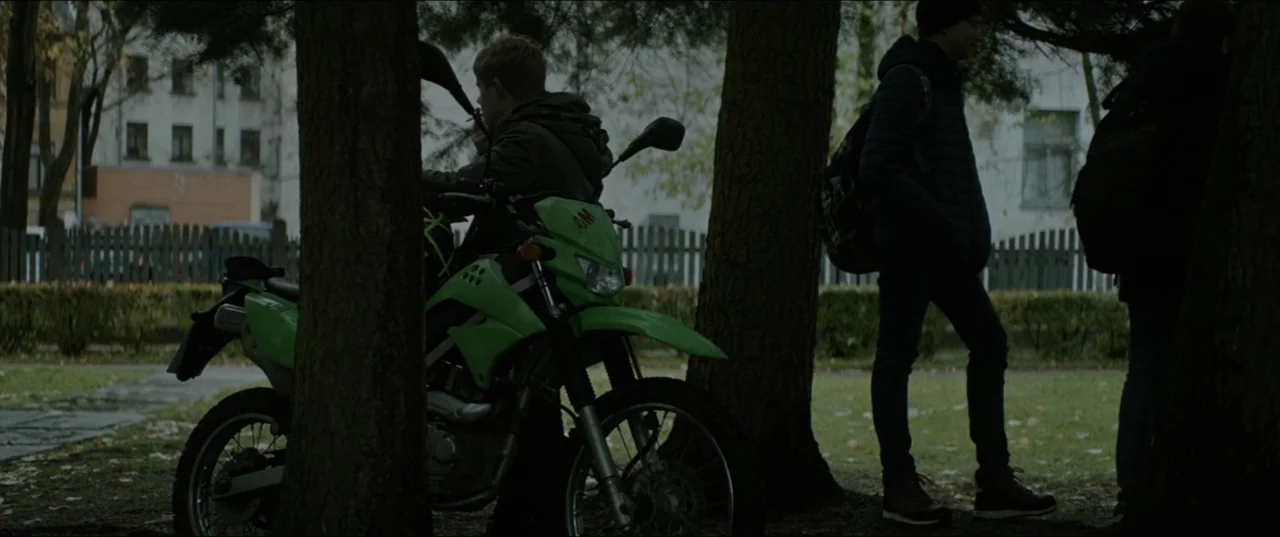 Dogs Don't Wear Pants (2019) movie still: medium — A park area with mature trees and a pale building in the background. A teenager sits on a bright…
