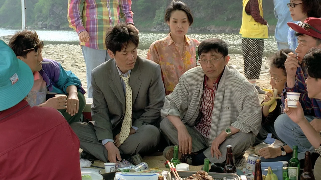 Peppermint Candy (1999) movie still: medium — A group of people sits on a straw mat near a riverbank. They are having a casual picnic with various…