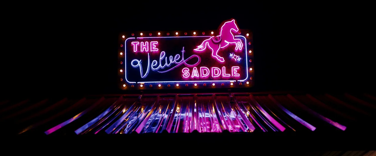 LaRoy, Texas (2023) movie still: wide — A glowing pink and blue neon sign reads The Velvet Saddle above a corrugated metal roof. Reflections from…