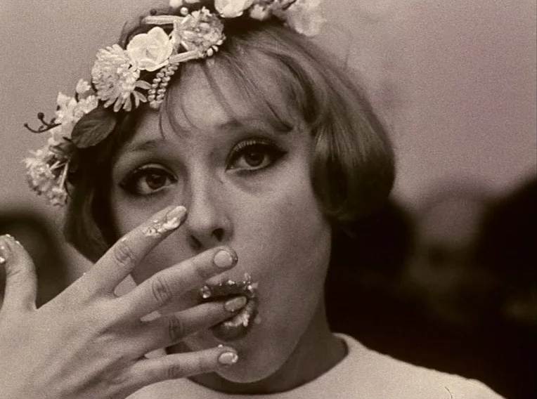 Daisies (1966) movie still: closeup — A close-up shot of a young woman wearing a floral headband. She is eating cake with her hand, leaving white…