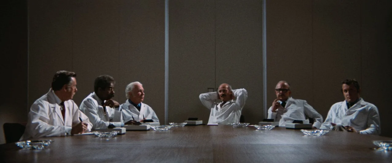 Westworld (1973) movie still: wide — A sterile boardroom features beige walls and a large, empty dark wood table. Six men in white coats sit around…