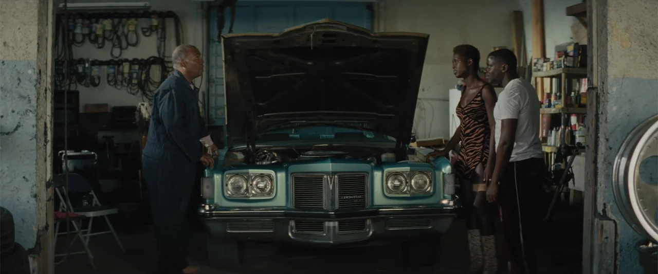 Queen & Slim (2019) movie still: fullbody — An auto repair shop interior filled with tools and shelves contains a turquoise car. A mechanic in blue…