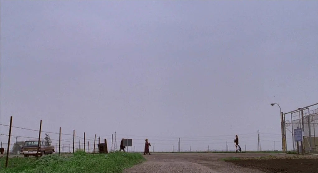 The Crossing Guard (1995) movie still: wide — An expansive overcast sky hangs over a flat, desolate dirt lot with distant power lines. Three people…