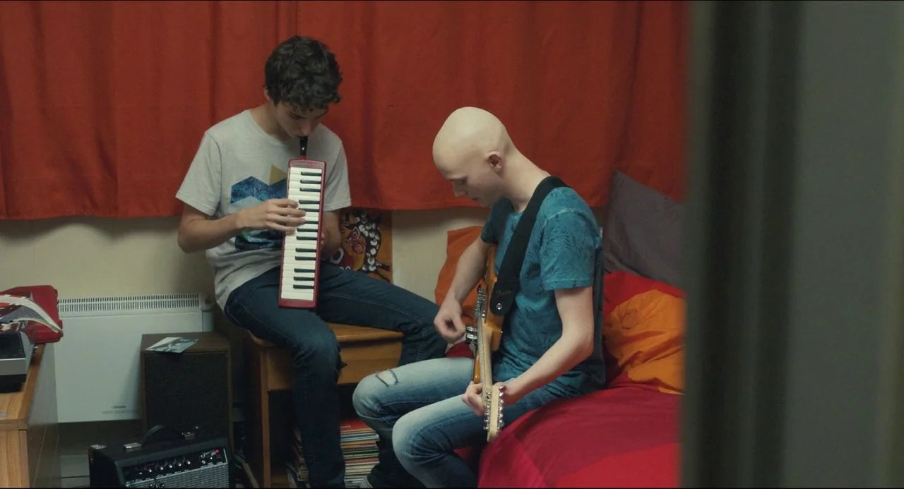 The Unknown Girl (2016) movie still: medium — A small room features orange curtains and a bed with red and orange bedding. One young man plays a red…