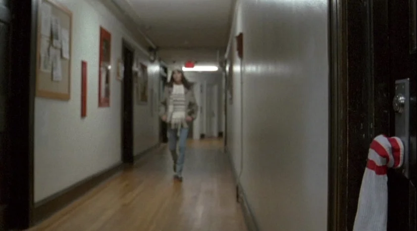 The House Of The Devil (2009) movie still: wide — A long, narrow apartment hallway with white walls and polished wooden floors. A woman walks toward…
