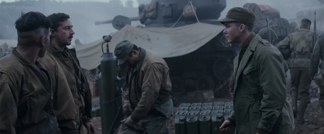 Fury (2014) movie still: medium — A military encampment featuring a large tank parked on muddy ground. Several soldiers in WWII-era uniforms stand…