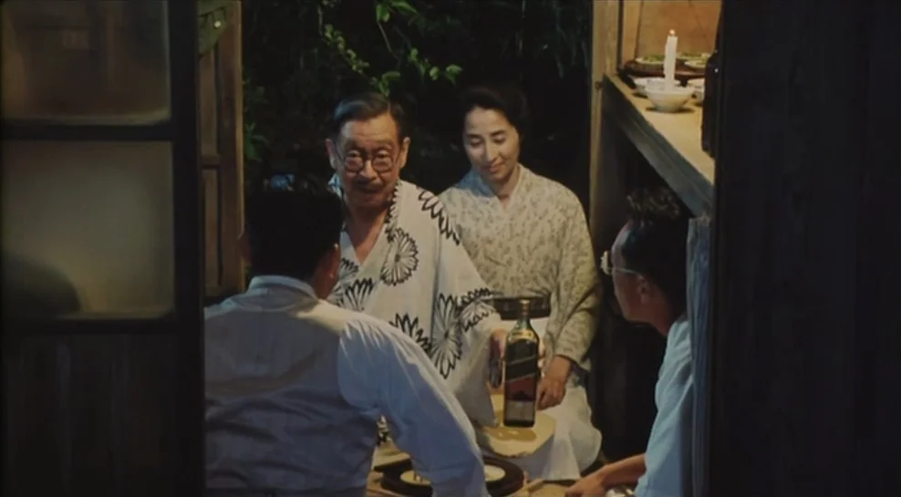 Madadayo (1993) movie still: medium — A wooden veranda opens onto a dark outdoor area with dense foliage at night. Four people sit together while an…