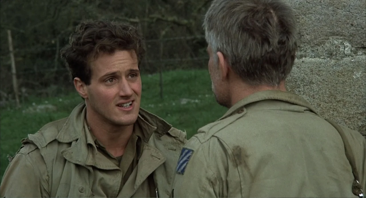Zone Troopers (1985) movie still: medium — Two soldiers in tan uniforms stand near a stone wall in an open field. The younger soldier faces his…