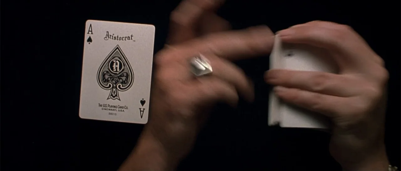Shade (2003) movie still: closeup — A dark, nondescript surface frames a single playing card next to a deck being shuffled. Hands manipulate a deck…