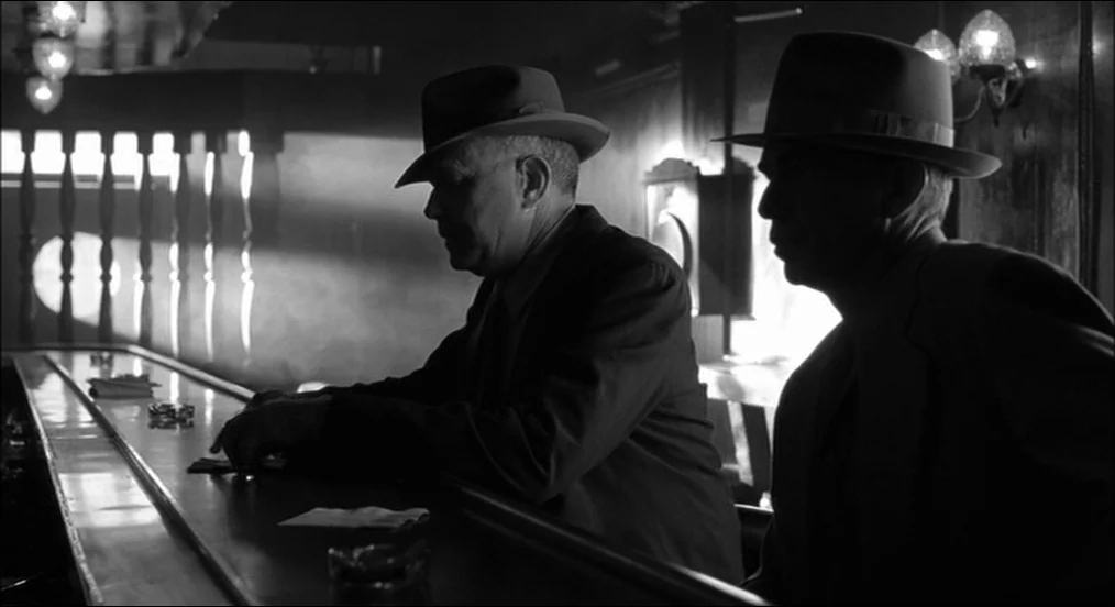 The Man Who Wasn’t There (2001) movie still: medium — A dimly lit 1940s style tavern features a long wooden bar counter. Two men in fedoras and…