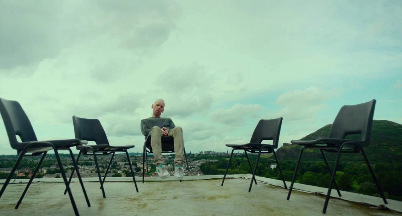 T2 Trainspotting (2017) movie still: wide — A flat concrete rooftop overlooks a small town and a large grassy hill. A solitary man sits on a black…