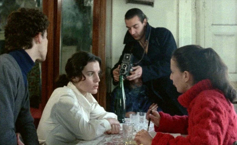 The Kingdom of Naples (1978) movie still: medium — A group of four young adults sit around a table in a dimly lit room. One man stands and aims a…