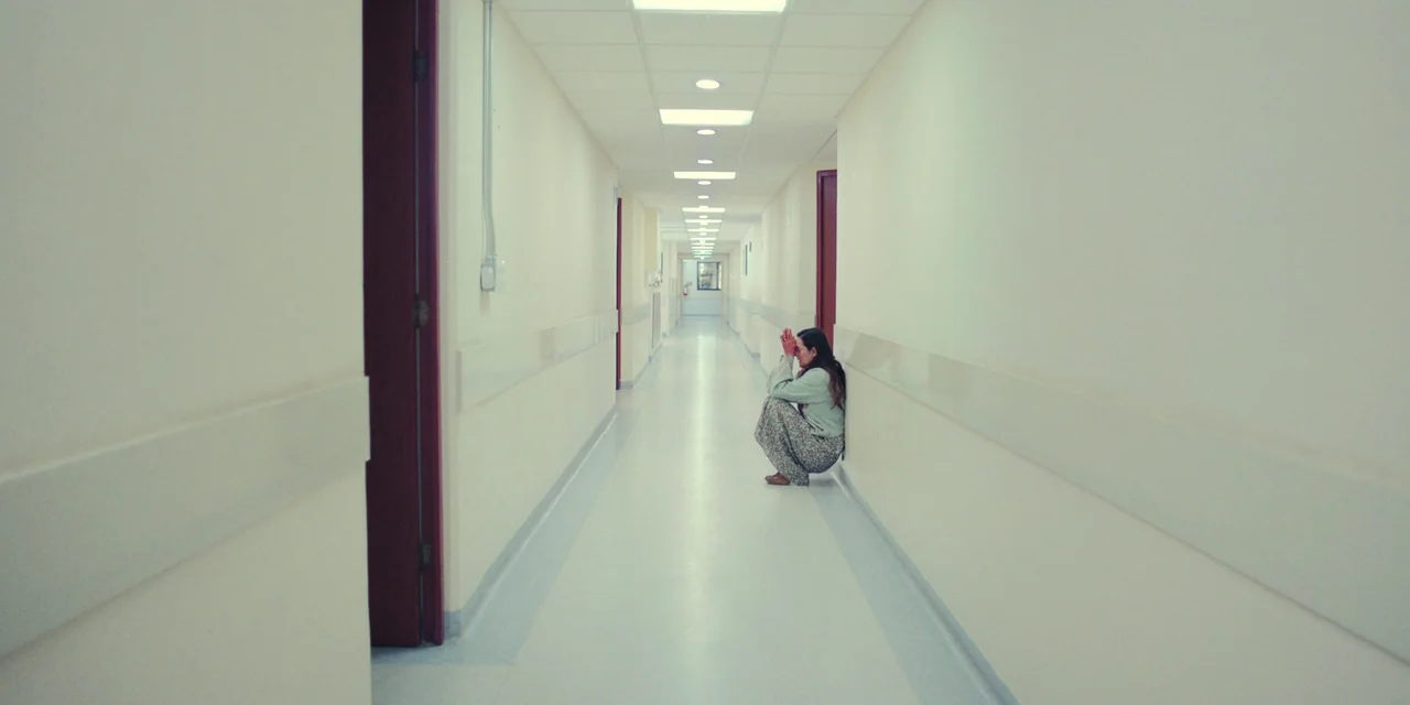 Silent Night (2023) movie still: wide — A long, sterile hospital hallway with white walls and a polished floor. A lone woman sits crouched against…