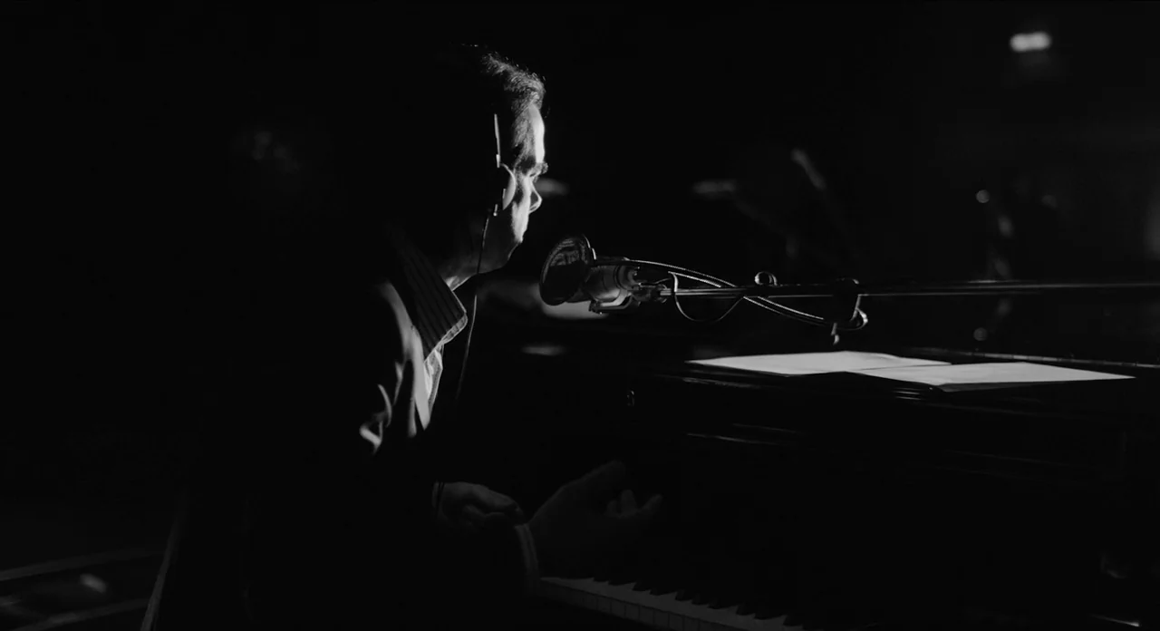 One More Time With Feeling movie still: medium — A dark, void-like recording studio contains only a piano and a microphone setup. A man in a suit…