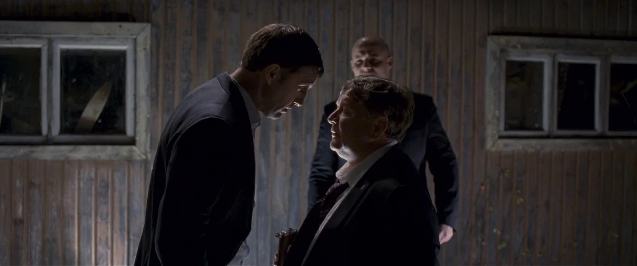 Leviathan (2014) movie still: medium — A dark wooden structure with two windows serves as the backdrop at night. Two men stand in close proximity…
