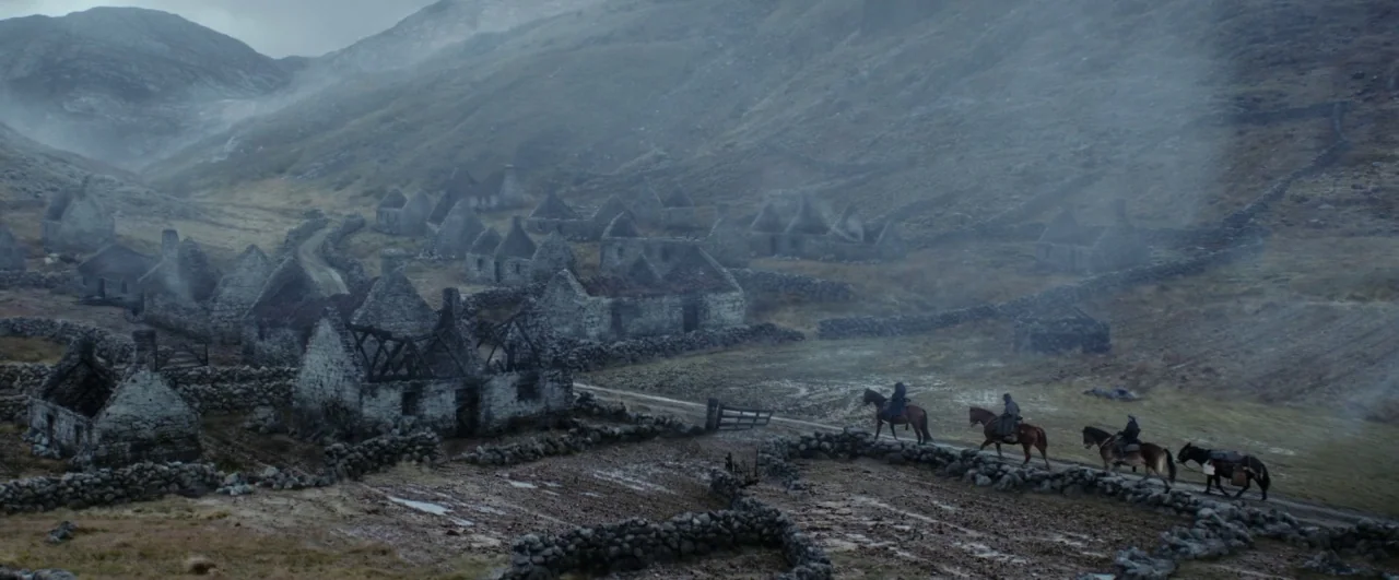 Black ’47 (2018) movie still: wide — A cluster of ruined stone cottages sits within a misty, mountain-flanked valley landscape. Four riders on…