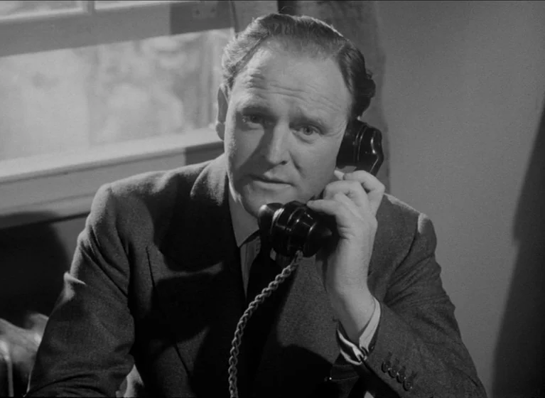 The Blue Lamp (1950) movie still: medium — A man sits at a desk in an office setting with a window behind him. He is holding a black rotary…