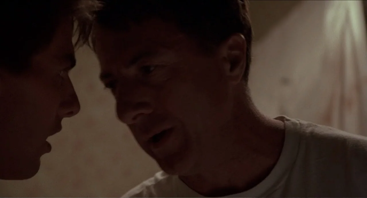 Rain Man (1988) movie still: closeup — A dimly lit room with muted wallpaper patterns provides the backdrop. Two men stand close together in…