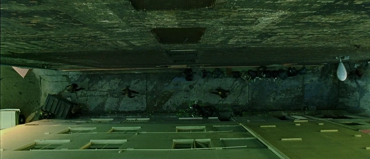 The Matrix (1999) movie still: wide — A narrow, paved urban passage between two brick buildings lined with refuse. Multiple black trash bags are…
