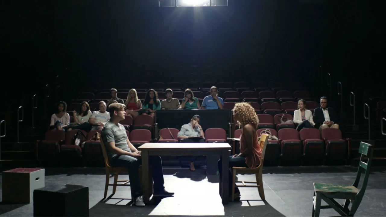 At Middleton (2013) movie still: wide — A dark auditorium with rows of empty red seats faces a central wooden table. A man and a woman sit at the…