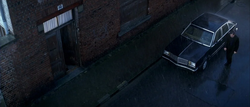 The Banishment (2007) movie still: wide — A dark brick building wall runs along a rainy road at night. A lone man stands next to a black sedan…