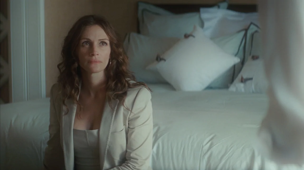 Duplicity (2009) movie still: medium — A modern hotel room features a large bed with white linens and pillows. A woman in a blazer sits on the edge…