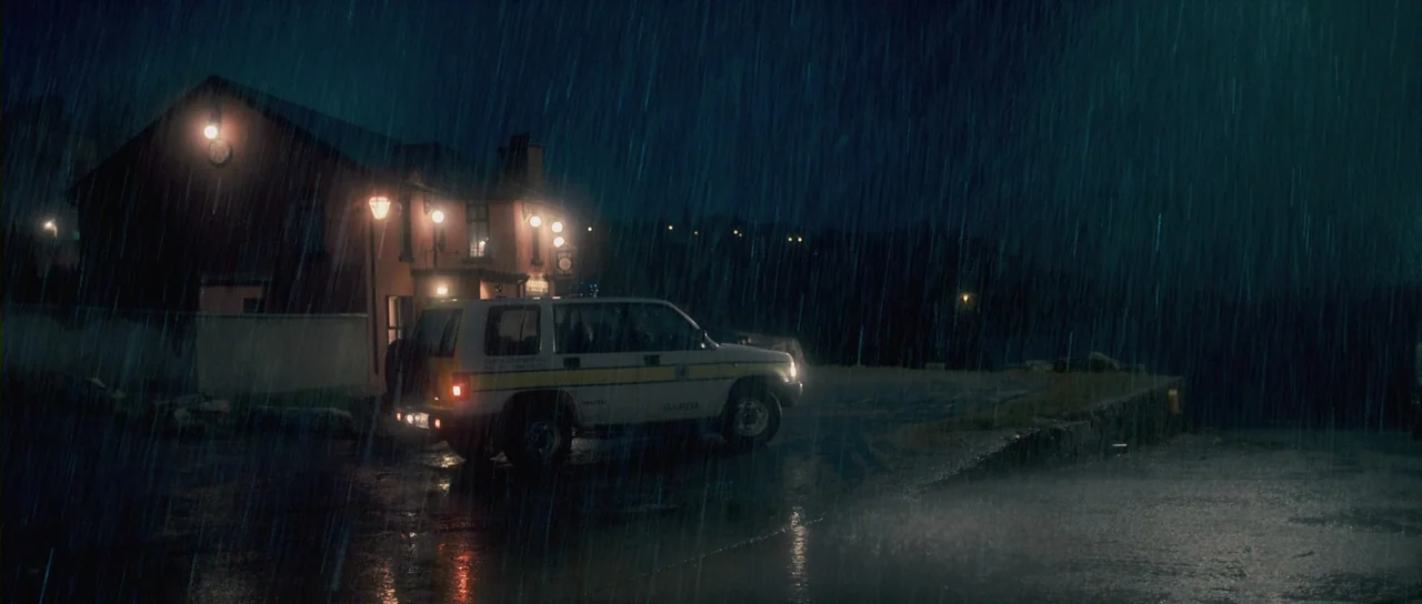 Grabbers (2012) movie still: wide — A rain-slicked dark road at night beside a glowing building exterior. A white police SUV is parked facing right…