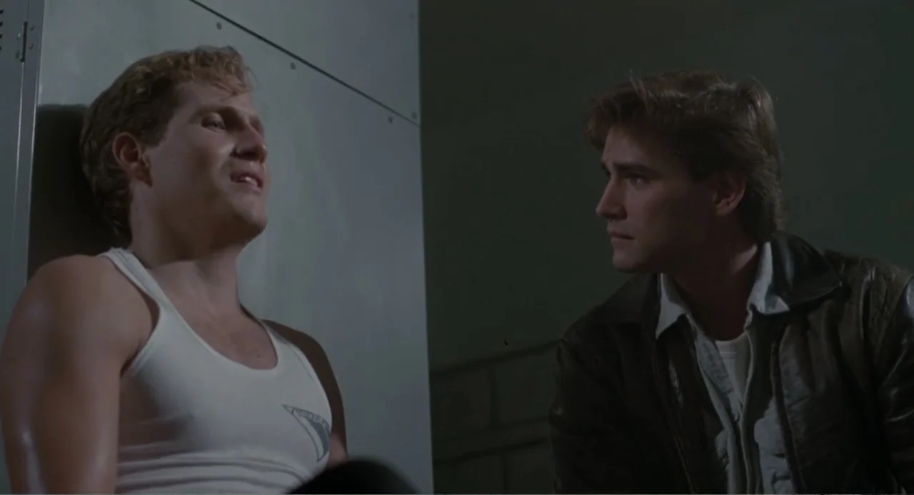 Dangerously Close (1986) movie still: medium — A dimly lit room features metal lockers along the wall in the background. Two young men are…