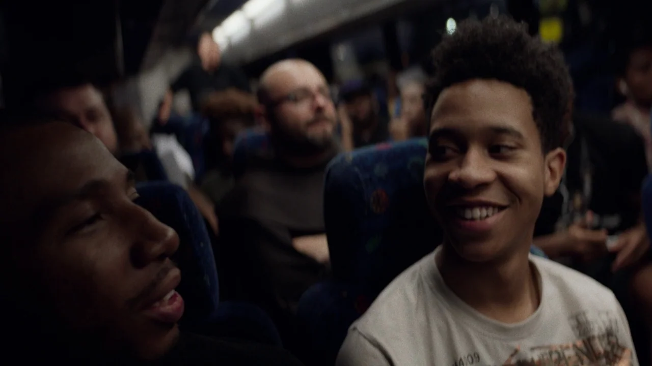Pass Over (2018) movie still: medium — The interior of a crowded bus is dimly lit with passengers visible in rows. Two men are seated together and…