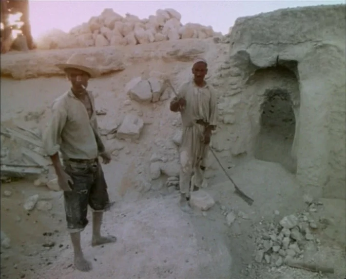 Fata Morgana (1971) movie still: fullbody — An arid archaeological excavation site features light sandy mounds and a small stone cave. Two men stand…