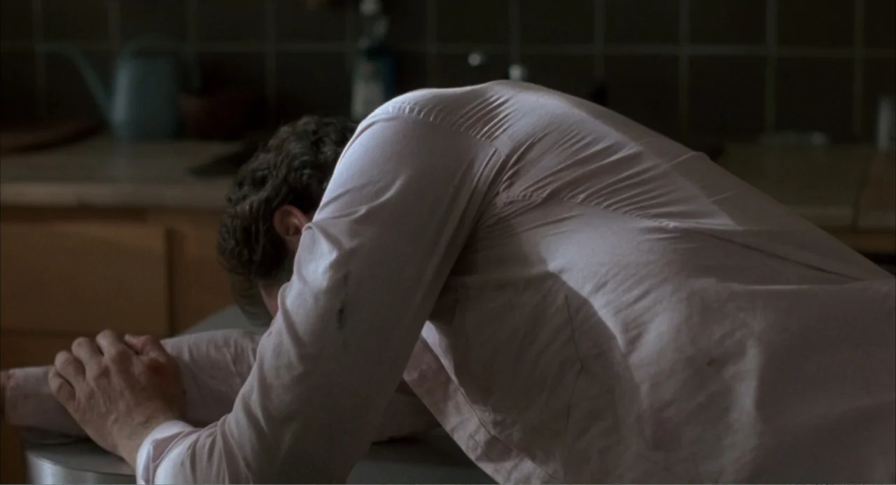 F/X (1986) movie still: medium — A dimly lit domestic kitchen features dark brown tiled walls and a countertop. A man wearing a stained button-down…