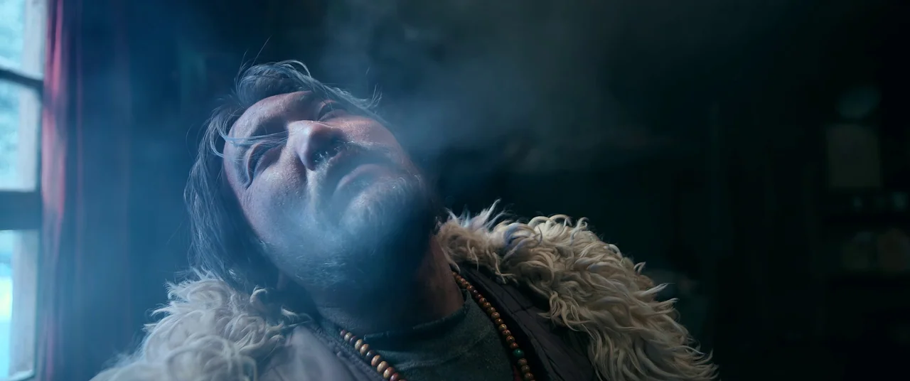 One And Four (2021) movie still: closeup — A man lies inside a dim cabin illuminated by blue light from a window. He exhales a thick plume of smoke…