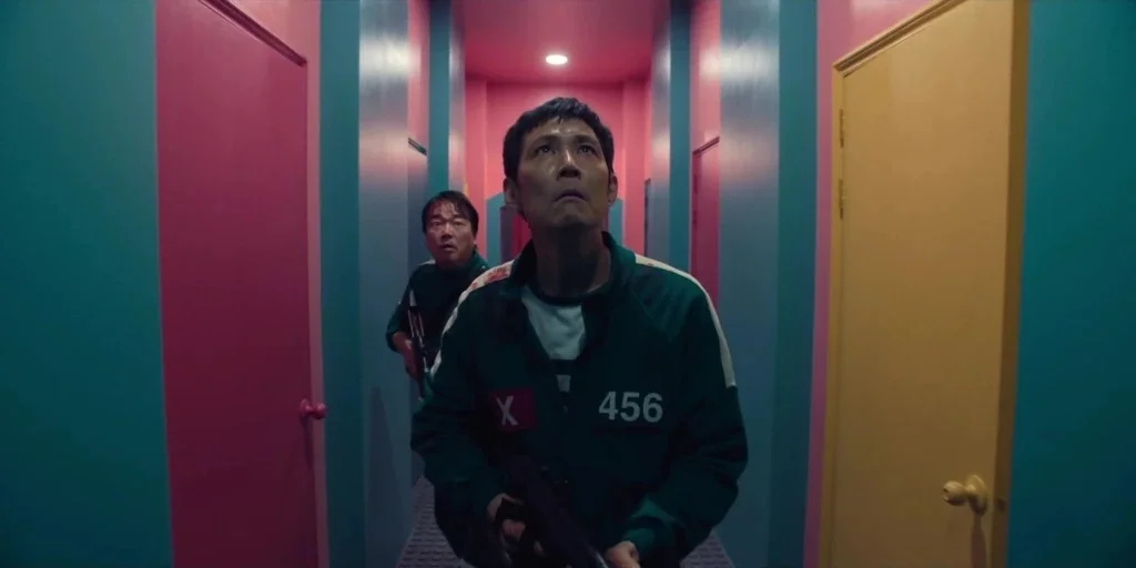 Squid Game (2024) movie still: medium — A windowless corridor features bright pink and teal vertical wall panels and yellow doors. Two men in green…