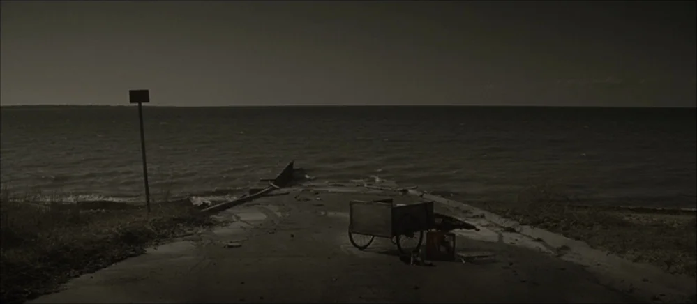 The Road (2009) movie still: establishing — A desolate paved pier extends into a dark, choppy body of water. A weathered cart and a red plastic…
