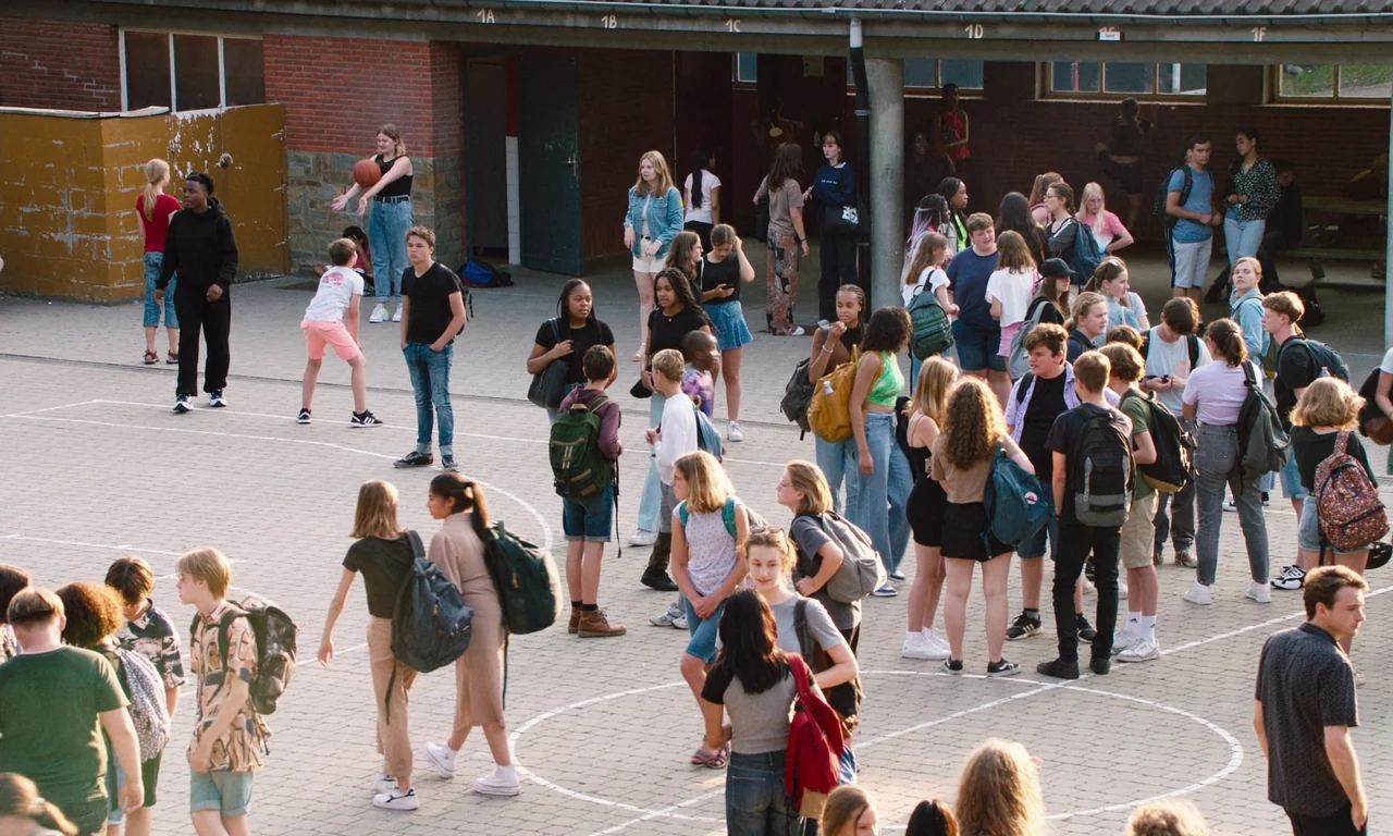 Close (2022) movie still: wide — A paved school courtyard is filled with a large crowd of diverse students milling about. Many teenagers stand in…