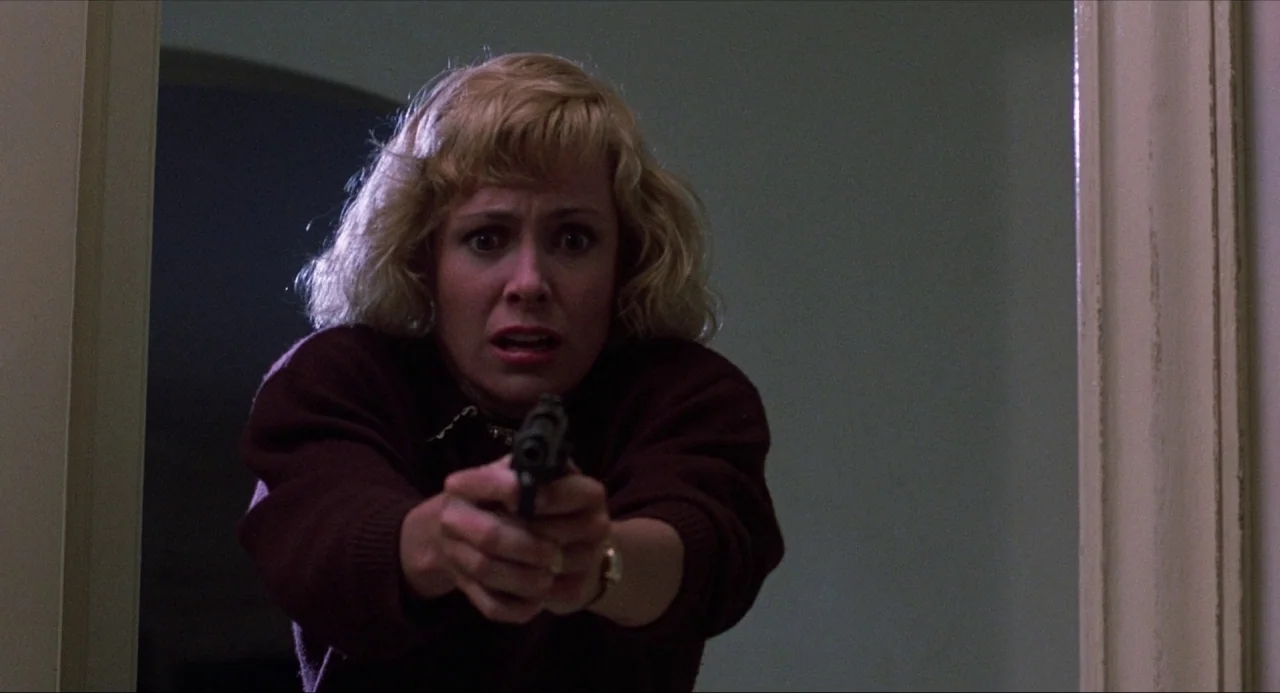 Child’s Play (1988) movie still: medium — A person stands in a dim doorway between two darkened spaces. A blonde woman in a burgundy sweater holds a…