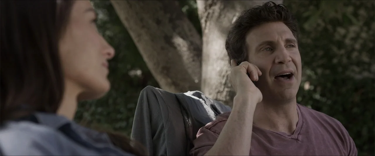 Heavenly Deposit (2019) movie still: medium — A sunny outdoor backyard area with trees visible in the soft-focused background. A man holds a phone…